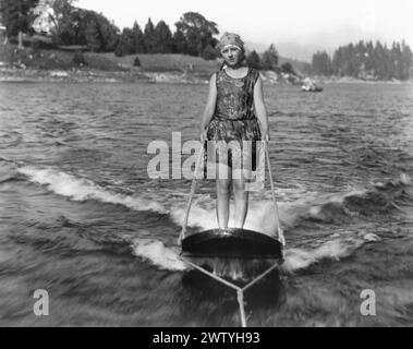 femme des années 1920 en maillot de bain et casquette chevauchant sur planche de surf derrière un bateau Banque D'Images