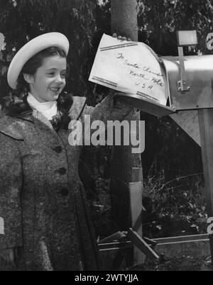 Fille dans un manteau d'hiver et chapeau à une boîte aux lettres postant une lettre au Père Noël Banque D'Images