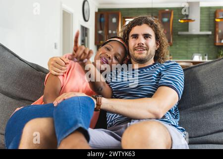 Un couple diversifié se détend sur le canapé à la maison, pointant vers quelque chose tout en regardant la télévision Banque D'Images