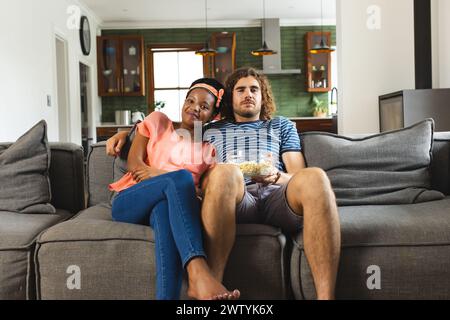 Un couple diversifié se détache sur un canapé gris à la maison, mange du pop-corn et regarde la télévision Banque D'Images