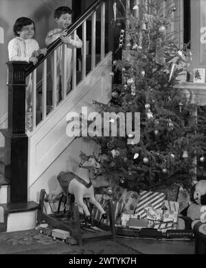 Jeune garçon et sa sœur en pyjama debout sur l'escalier à la rampe regardant vers le bas sur un arbre de Noël chargé de jouets et de cadeaux le matin de Noël Banque D'Images