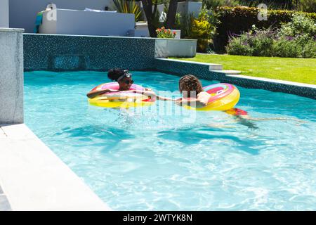 Un couple diversifié profite d'une journée ensoleillée à la piscine à la maison, flottant sur des anneaux colorés avec espace de copie Banque D'Images