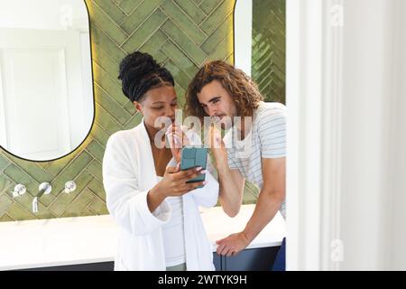 Un couple diversifié examine un smartphone ensemble tout en se brossant les dents dans une salle de bain Banque D'Images