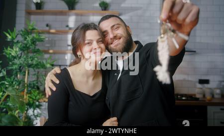 Jeune couple nouvellement marié assis dans la cuisine sourire regarder la caméra mari montrer des clés de la nouvelle maison. Fiers propriétaires, appartement loué, prêt hypothécaire c Banque D'Images