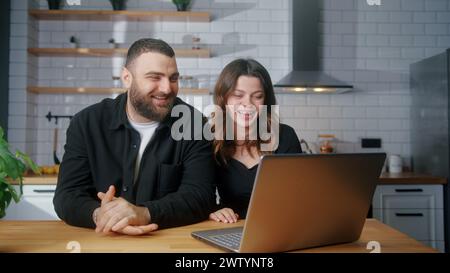 Heureux couple assis dans la cuisine parler distance appel vidéo regardant la webcam de l'ordinateur portable. Parler en ligne avec des amis et des parents Banque D'Images