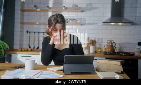 Jeune femme assise dans la cuisine étudiant avec sa tablette et prend des notes dans un cahier. Femme réfléchie vient à l'esprit avec une nouvelle idée, étude conc Banque D'Images