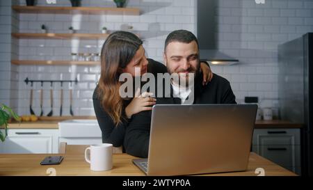 Jeune homme freelance assis dans la cuisine travaillant sur ordinateur portable sa petite amie vient de derrière câlins, souriant Banque D'Images