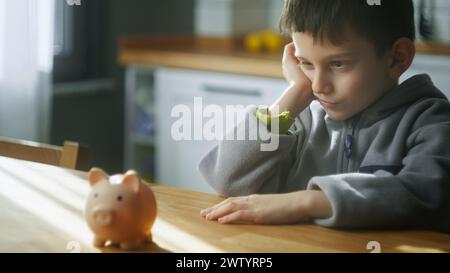 Boy mignon ennuyé assis dans la cuisine en pensant et en regardant une tirelire. Concept d'enseignement de la littératie financière, planification budgétaire Banque D'Images