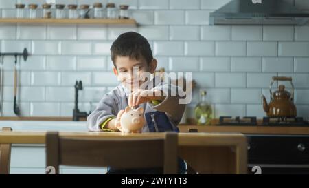 Garçon souriant assis dans la cuisine mettant des pièces dans une tirelire. L'enfant économise de l'argent et investit dans l'avenir. Concept de l'enseignement de la littérature financière Banque D'Images