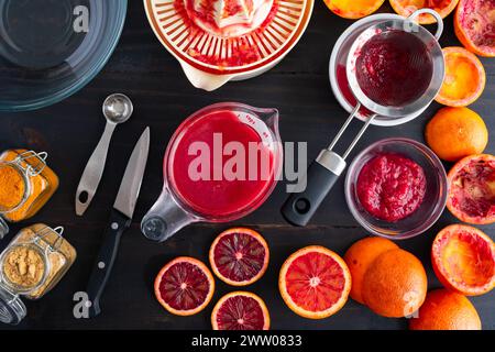 Jus d'orange sanguine dans une tasse à mesurer sur une table en bois : oranges sanguines juxtaposées et coupées en deux avec centrifugeuse électrique, passoire et autres outils de cuisine Banque D'Images