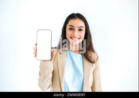 Excité heureuse femme brune indienne ou arabe en costume élégant, montrant un téléphone intelligent avec un écran de maquette blanc vide pour la publicité, debout sur fond blanc isolé, regardant la caméra, souriant Banque D'Images