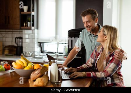 Un couple joyeux profite d'un moment léger dans leur cuisine ensoleillée, travaillant sur ordinateur portable entouré d'un petit déjeuner sain Banque D'Images