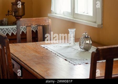 Une vieille table en chêne et trois chaises se tiennent dans un intérieur antique par une fenêtre. Une nappe blanche, un vase en cristal et un grand réveil rétro sont à votre disposition Banque D'Images