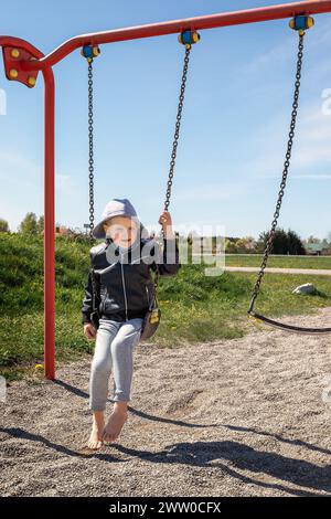 Un petit garçon, pieds nus, sans dents dans une veste en cuir noir est assis sur une balançoire de chaîne. Banque D'Images