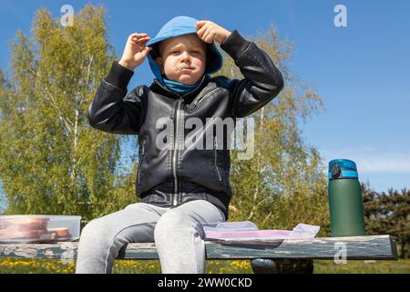 Le garçon est en train de déjeuner dans la nature sur un banc. Bon appétit à l'air frais, il colle la bouche pleine et s'assied avec des joues bouffantes. Banque D'Images