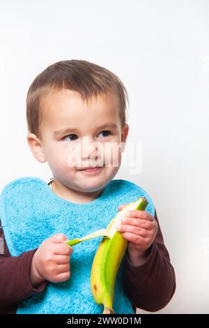 Photographie verticale d'un garçon avec un bavoir bleu épluchant une banane pour la manger. Fond blanc. Isolé. Banque D'Images