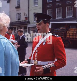 Années 1960, historique, une garde irlandaise en uniforme en tunique et chapeau de fourrage en présence pour « The Most Noble Order of the Jarter », un événement cérémonial spécial qui a lieu au château de Windsor, Berkshire, Angleterre, Royaume-Uni, donnant un dépliant d'ordre de service à une invitée. Formés en 1900 par la reine Victoria, les Irish Guards sont un régiment de l'Arrmy britannique et une partie de leurs fonctions est de garder la famille royale britannique et de prendre part aux cérémonies traditionnelles. Banque D'Images