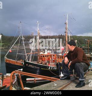 Années 1960, historique, un pêcheur assis sur des bateaux de pêche de quai, Stornoway, île de Lewis, îles occidentales, Écosse, ROYAUME-UNI. Stornoway a été le centre de l'industrie britannique du hareng pendant des siècles, avec l'apogée de l'industrie entre 1900 et la 1ère Guerre mondiale. Alors que les habitudes alimentaires nationales ont vu la pêche au hareng décliner, l'industrie reste économiquement importante pour l'île. Banque D'Images