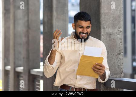 Un homme barbu joyeux se tient dehors, souriant largement en lisant une lettre dans une enveloppe jaune. L'image capture un sentiment de joie, de surprise et d'émotion positive, idéal pour les concepts de bonne nouvelle, de communication et de bonheur. Banque D'Images