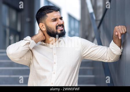 Photo en gros plan d'un jeune homme arabe fatigué debout près d'un immeuble de bureaux, appuyant sa main sur le mur et massant son cou, souffrant et grimaçant de douleur et de tension. Banque D'Images