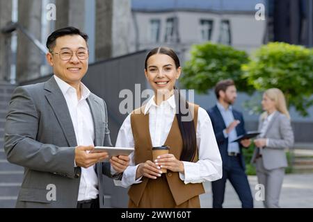 Deux collègues d'affaires joyeux avec tablette numérique debout à l'extérieur, avec d'autres membres de l'équipe en arrière-plan. Banque D'Images