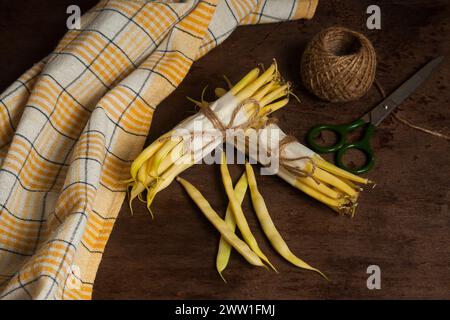 Deux bouquets et plusieurs gousses de haricot jaune cru avec serviette de cuisine jaune, ciseaux et boule de fil sur fond de bois. Haricots ou Banque D'Images