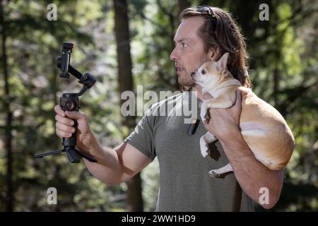 Homme utilisant un cardan de téléphone pour filmer lui-même et un chihuahua qu'il tient. Le film se passe dans une forêt naturelle. Banque D'Images