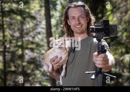 Homme utilisant un cardan de téléphone pour filmer lui-même et un chihuahua qu'il tient. Le film se passe dans une forêt naturelle. Banque D'Images