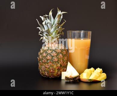 Un mélange de jus de fruits dans un grand verre à facettes sur fond noir, à côté de morceaux d'ananas mûrs et de mangue. Gros plan. Banque D'Images