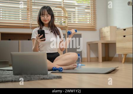 Une jeune femme asiatique heureuse en vêtements de sport fait de l'exercice avec un haltère sur un tapis de yoga dans le salon tout en discutant avec ses amis sur son smartpho Banque D'Images