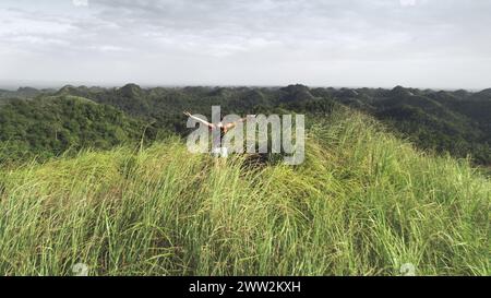 Sommet de montagne aérienne : femme lever les mains au sommet de l'herbe haute des collines des Philippines, Asie. Paysage épique avec fille de vue arrière. Majestueuse nature asiatique avec admirez les touristes. Prise de vue cinématographique par drone Freedom Banque D'Images