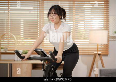 Une jeune femme asiatique forte et en forme dans les vêtements de sport fait de l'exercice sur un vélo de fitness, faisant du cardio d'intérieur à la maison. personnes et concepts de sport d'intérieur Banque D'Images