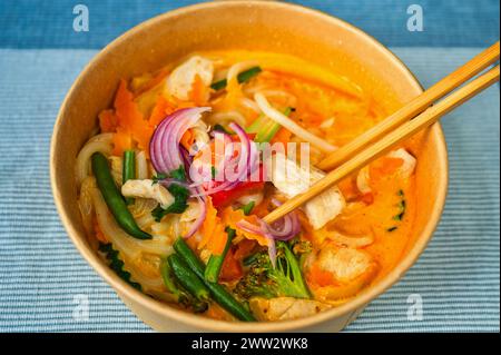 Curry rouge épicé avec légumes, viande de poulet et lait de cacao dans un bol et baguettes sur fond bleu, gros plan. Cuisine asiatique. Banque D'Images