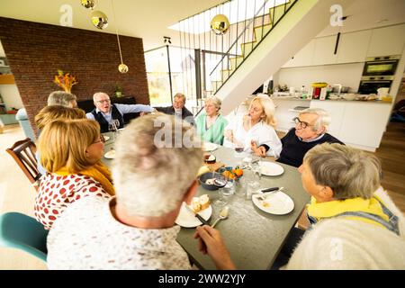 Un rassemblement familial animé autour d'une table de dîner, immergé dans la conversation dans une maison moderne. La scène se déroule avec une cuisine spacieuse en toile de fond, de grandes fenêtres et un escalier élégant, ajoutant une touche de sophistication au repas familys. Souper familial joyeux dans une maison moderne avec conversations. Photo de haute qualité Banque D'Images