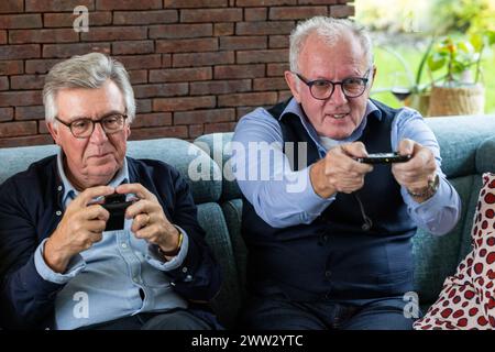 Deux hommes âgés font preuve de concentration et de plaisir tout en jouant à des jeux vidéo, assis confortablement sur un canapé. Un mur de briques offre une toile de fond chaleureuse, soulignant le passe-temps moderne dans un environnement accueillant. Les hommes seniors sont plongés dans les jeux vidéo sur le canapé. Photo de haute qualité Banque D'Images