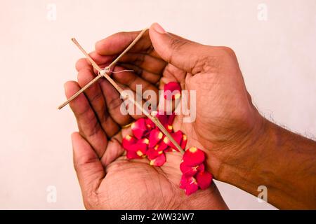 Une photo en gros plan de mains douces berçant une simple croix en bois, ornée de pétales de rose rouge éparpillés Banque D'Images