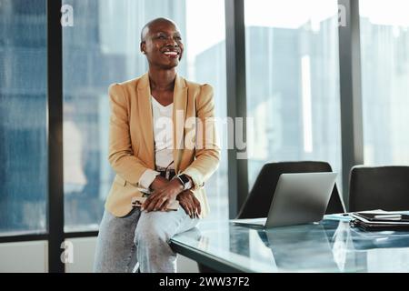 Femme d'affaires africaine confiante dans un bureau, assise à une table. Elle est heureuse et réussie, souriante tout en regardant loin. Son professionnalisme et orl Banque D'Images