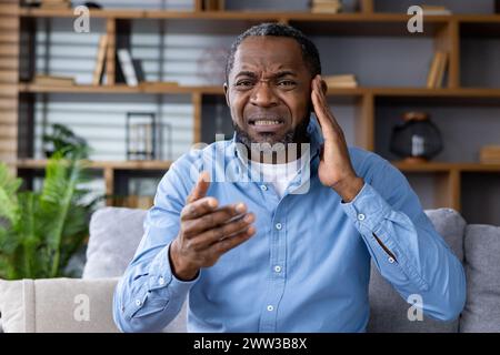 Homme mature avec une expression douloureuse touchant son oreille tout en discutant des symptômes avec un médecin lors d'un appel de consultation en ligne. Banque D'Images