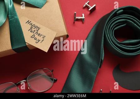 Carte avec phrase joyeuse fête des pères, boîte cadeau et accessoires pour hommes sur fond rouge, plat Banque D'Images