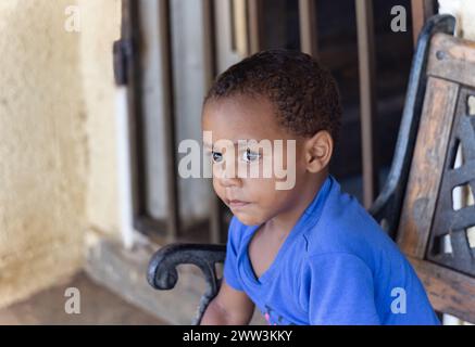 garçon africain avec un regard sérieux, assis sur un banc, devant la maison dans le township, en fin d'après-midi, Banque D'Images