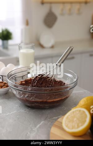 Fouet en métal, crème au chocolat dans un bol et différents produits sur une table en marbre gris à l'intérieur Banque D'Images