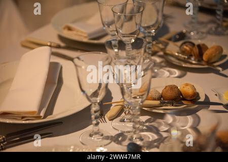 Gros plan sur un décor raffiné, avec des verres cristallins, de la vaisselle blanche élégante et des serviettes soigneusement pliées. L'arrangement invitant est complété par une sélection de petits pains fraîchement cuits, en attente de l'arrivée des invités. Table raffinée avec verres en cristal et vaisselle élégante. Photo de haute qualité Banque D'Images