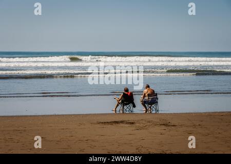 Un homme et une femme assis dans des chaises de pelouse sur la plage près du bord de l'eau regardant les vagues et la marée rouler. Banque D'Images