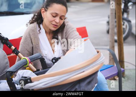 Portrait en gros plan d'une jeune jolie mère assise sur un banc de parc urbain coloré dans les couleurs du drapeau LGBT arc-en-ciel, avec son bébé nouveau-né dormant dans le b. Banque D'Images