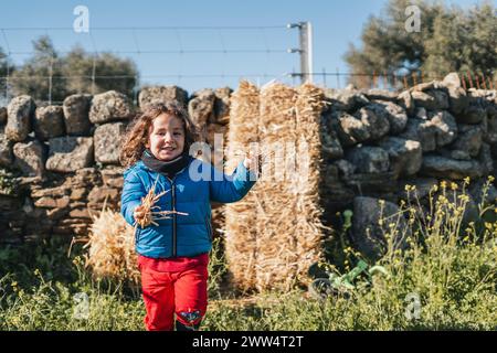 Un jeune garçon joue dans un champ avec des balles de foin et un mur de pierre. Elle tient des bâtons dans ses mains et elle s'amuse Banque D'Images