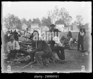 Protestation de l'armée de Coxey, 1894. Campement alors qu'ils marchent vers Washington, DC. Des hommes assis sur du bois et mangeant de la nourriture au camp de protestation de l'armée de Coxey, certains regardent la caméra, des maisons sont vues en arrière-plan, Banque D'Images