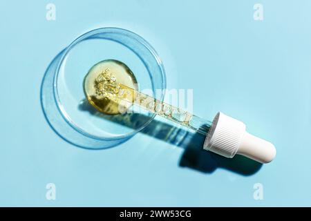 Boîte de Petri avec pipette et échantillon de sérum cosmétique doré pour le visage sur fond bleu, vue de dessus. Recherche en sciences biologiques, rétinol ou concept de soins de la peau peptidique Banque D'Images