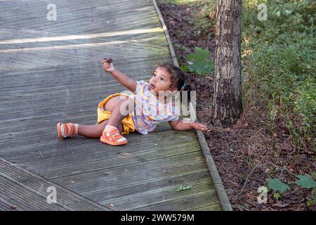 Une jeune fille de race mixte est assise sur une passerelle en bois, tenant une pomme de pin, avec des arbres et de la verdure autour, évoquant Banque D'Images