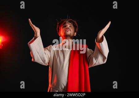 Homme en robe de Jésus et couronne d'épines étirant les mains avec la lumière rouge sur fond noir Banque D'Images