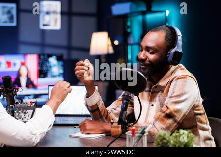 Homme noir souriant faisant la bosse de poing avec le présentateur afro-américain avant de commencer la diffusion de radio en direct. Animateur masculin joyeux saluant un invité lors d'une session d'enregistrement d'un talk-show en ligne. Banque D'Images